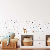 A minimalist childrens room has a white wall with My Stores Boho Polka Dots Stickers. A wicker basket holding a plush whale is next to a white cabinet with two woven baskets and whimsical mushroom decorations on top.