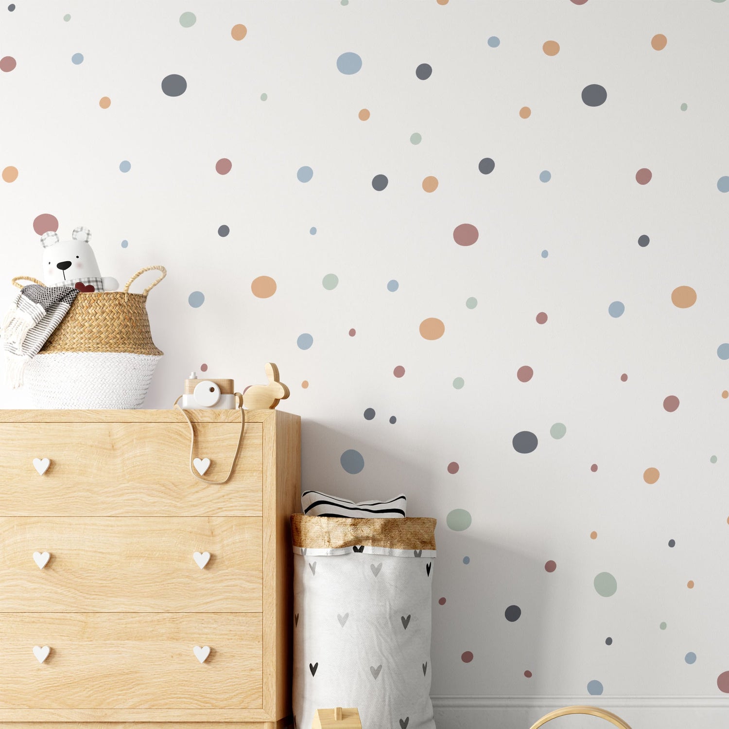 A nursery room includes a wooden dresser with heart-shaped handles, a plush bear, and a wicker basket. The wall displays My Stores Boho Polka Dots Stickers in various colors, adding charm beside the minimalist laundry basket.