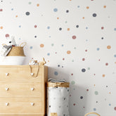 A nursery room includes a wooden dresser with heart-shaped handles, a plush bear, and a wicker basket. The wall displays My Stores Boho Polka Dots Stickers in various colors, adding charm beside the minimalist laundry basket.