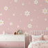 A childs room is decorated with My Stores Daisy Wall Decals and pink-white floral wallpaper. It includes a white metal bed with pink bedding, a pink and white dresser with mushroom ornaments, and small wooden houses on top—a lovely nursery haven.