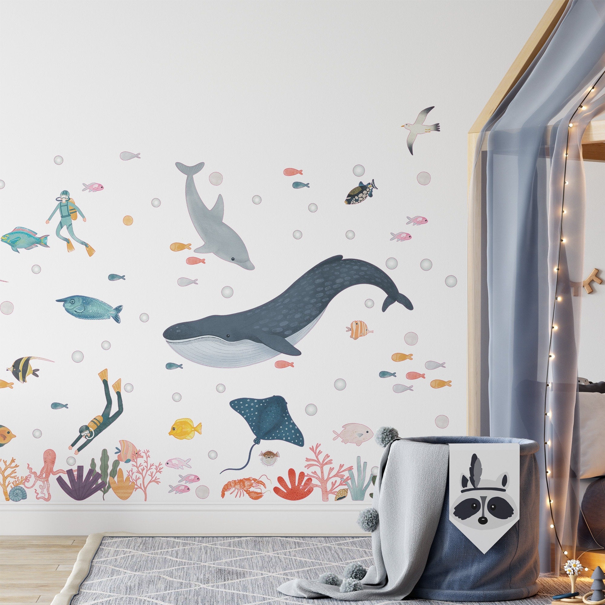 A childrens room with My Stores Sea Animals Stickers displays a whale, dolphin, and various fish. A cozy nook illuminated with string lights and complemented by a gray fabric basket featuring a raccoon face adds playfulness to the ocean-themed decor.