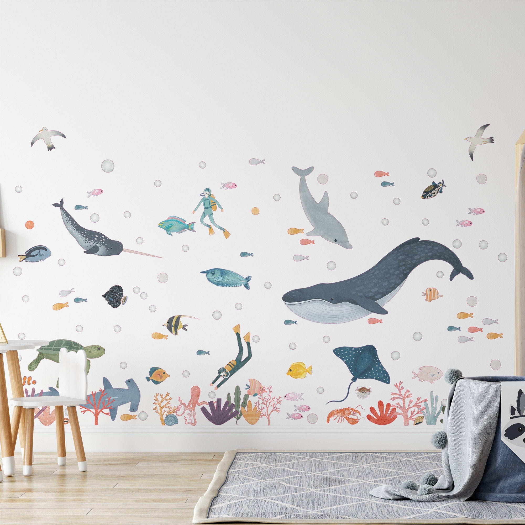 A childrens room decorated with My Stores Sea Animals Stickers, showcasing whales, dolphins, fish, coral, and divers. A small chair and round rug complete the playful underwater theme.