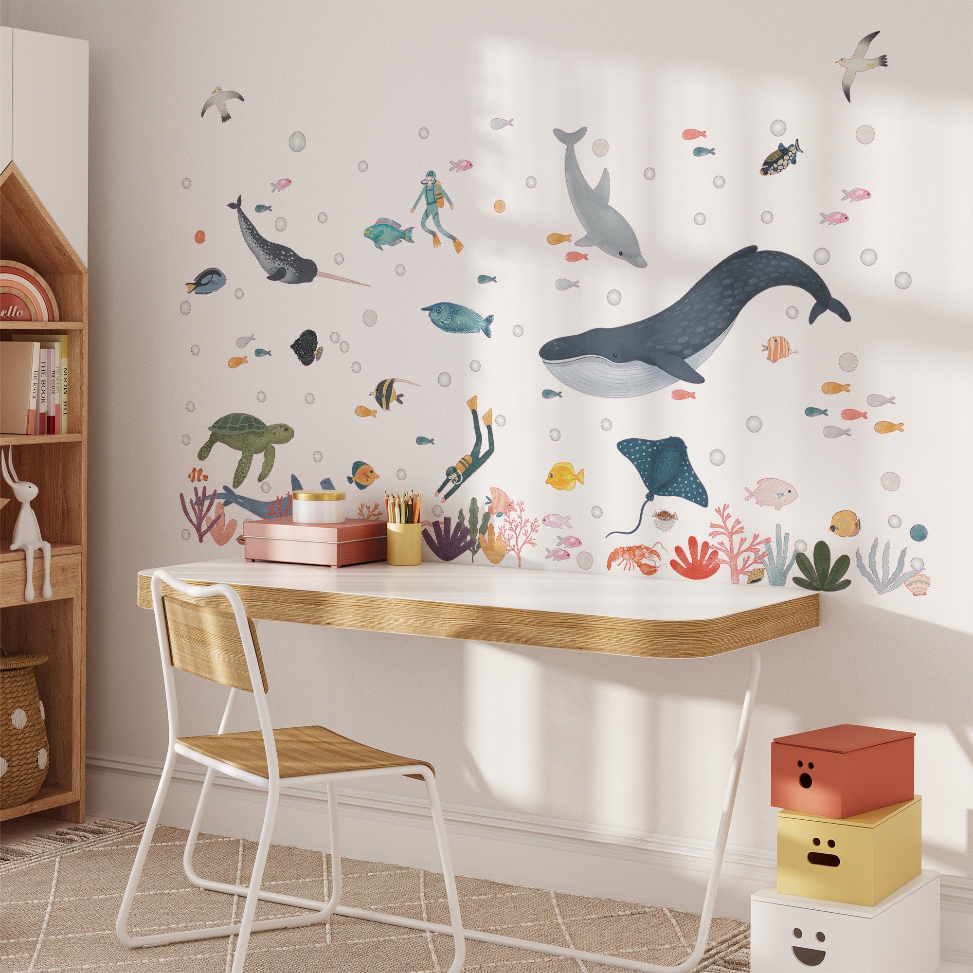 A childs room adorned with My Stores Sea Animals Stickers, featuring whales, turtles, and fish, has a wooden desk and chair. Sunlight illuminates the space with a small wooden shelf and playful storage boxes.