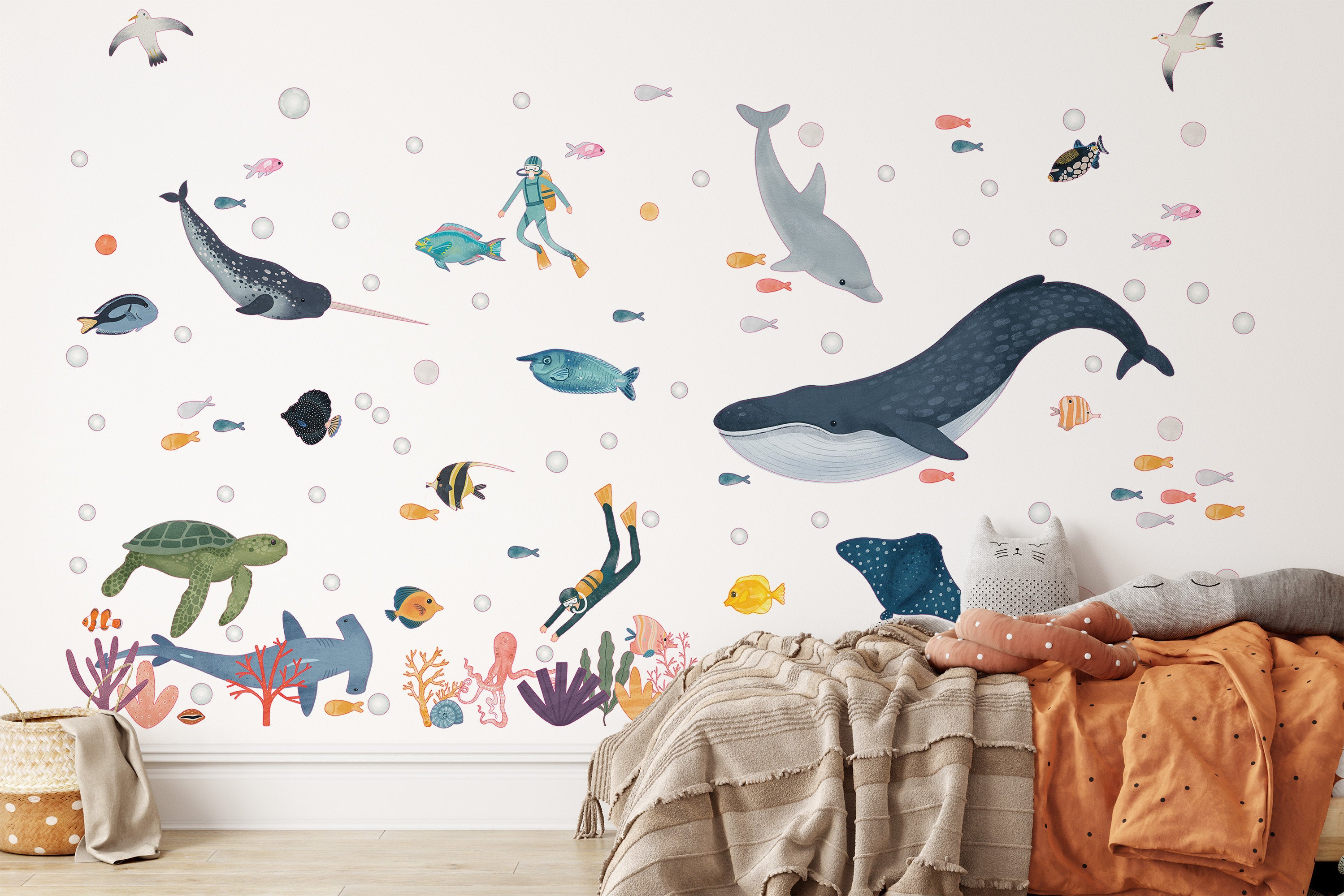 A vibrant underwater scene featuring My Stores Sea Animals Stickers, including fish, a whale, dolphins, seabirds, a diver, turtles, and coral reefs decorates the wall. Cozy bedding with cushions shaped like a cat face appears in the foreground.