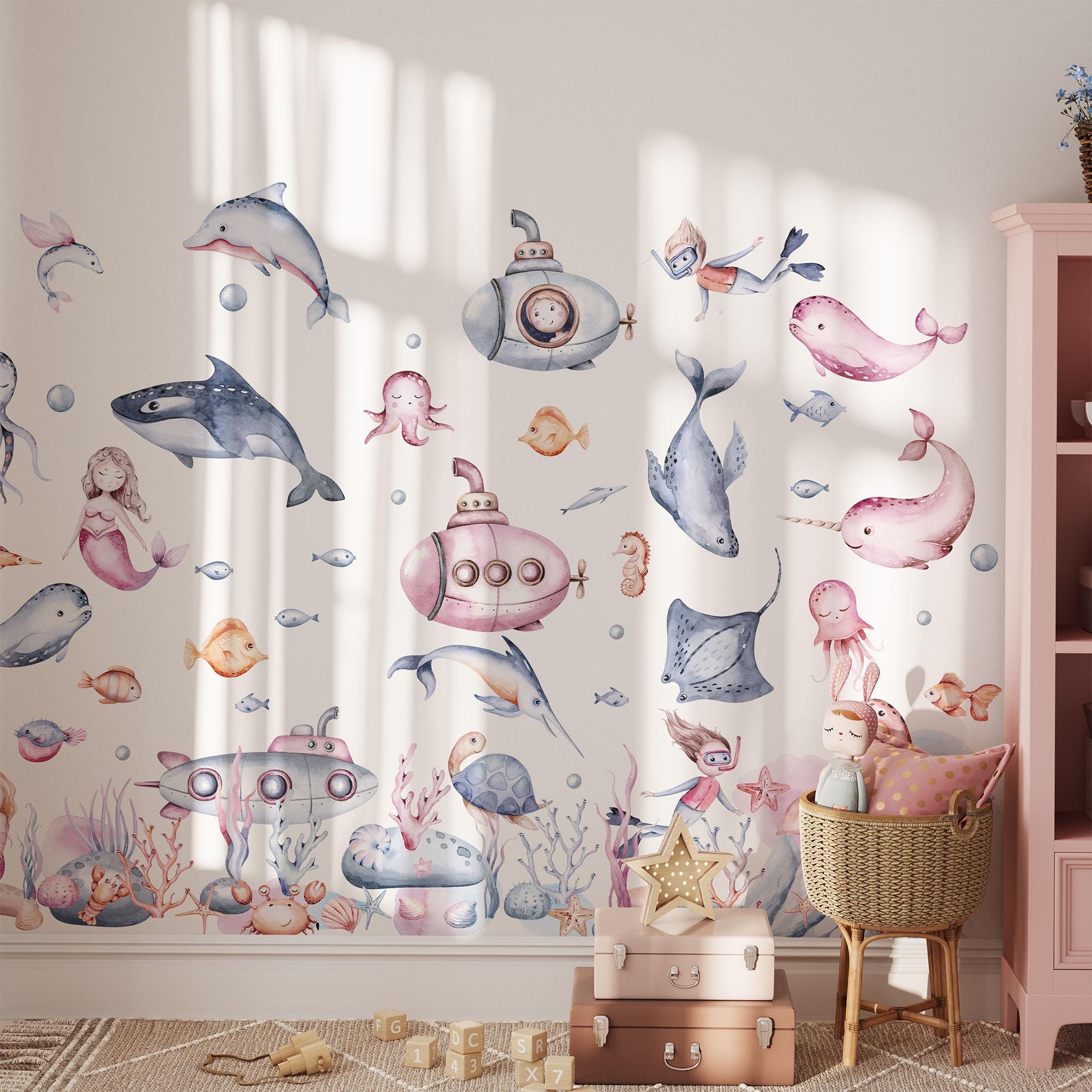A whimsical childrens room is adorned with My Stores Under The Sea Nursery Wall Stickers, featuring submarines, dolphins, mermaids, and fish. A basket of soft toys and a shelf of books enhance the playful decor. These removable decals spark endless undersea adventures.