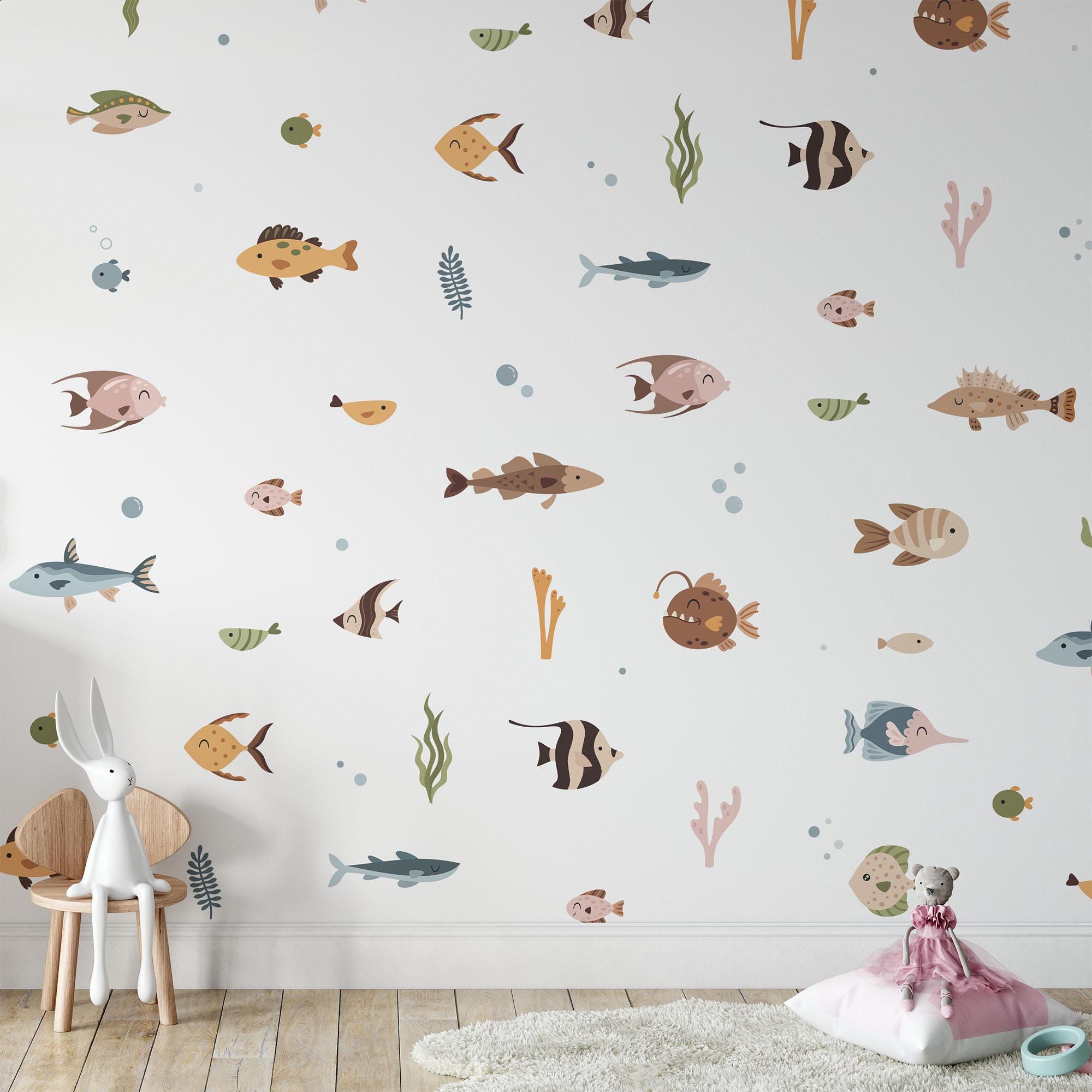 A childrens room features a white wall decorated with My Stores Ocean Fish Stickers showcasing cartoon fish and seaweed. A small chair holds a stuffed rabbit in the corner, and on the wooden floor rests a cushion with another cuddly toy, enhancing the ocean life theme.