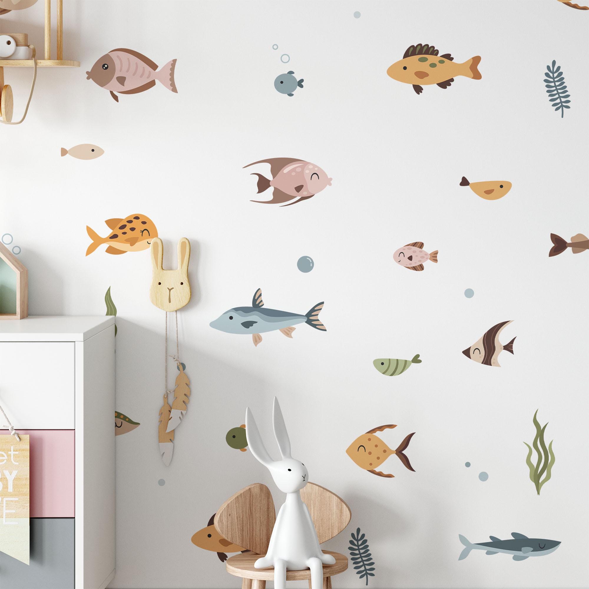A childrens room is decorated with My Stores Ocean Fish Stickers, featuring cartoon fish and sea animals. A small wooden chair with a gray bunny toy sits next to a white cabinet, enhancing the cozy, ocean-themed atmosphere.