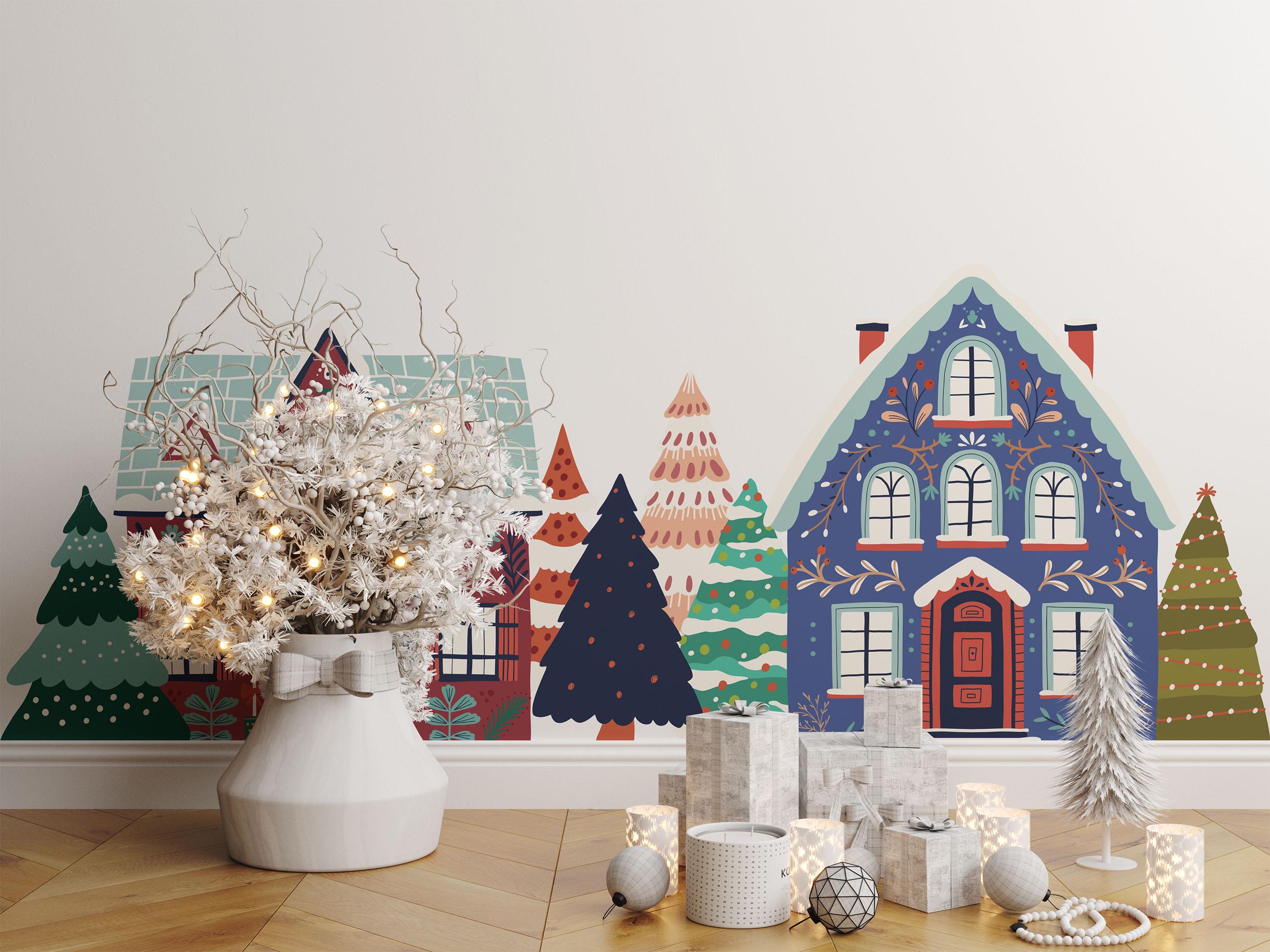 A festive scene features a Christmas tree in a white vase, paper houses and trees, with My Stores Festive Houses Wall Stickers adding colorful, whimsical holiday art. Candle holders and a small silver tree adorn the wooden floor.