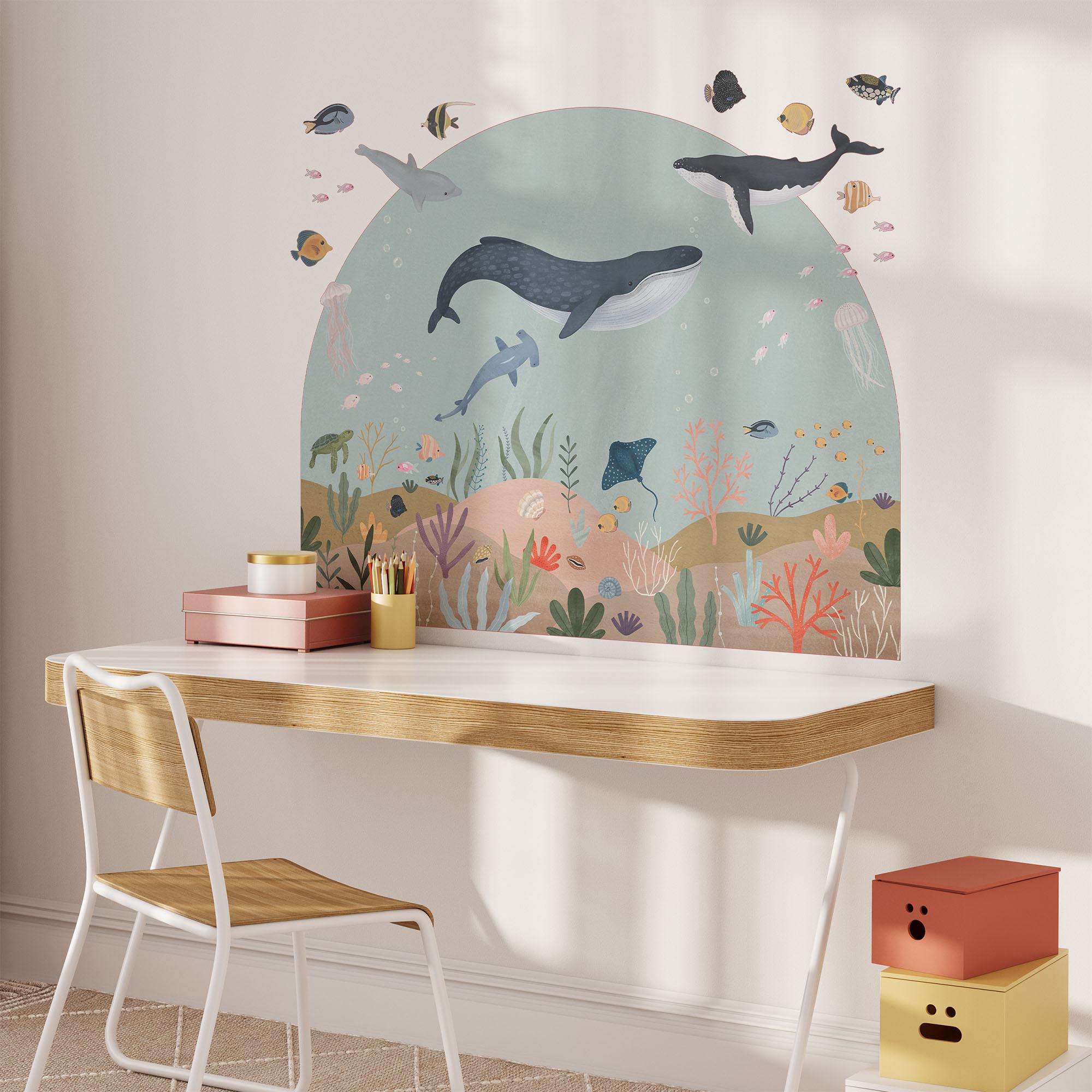 A childrens room with a light wooden desk and white chair features the Sea Animals Arch Wall Sticker by My Store, showcasing colorful whales, fish, coral, and sea plants. On the desk are stacked books, two pencils in a holder, and a surprised face storage box.