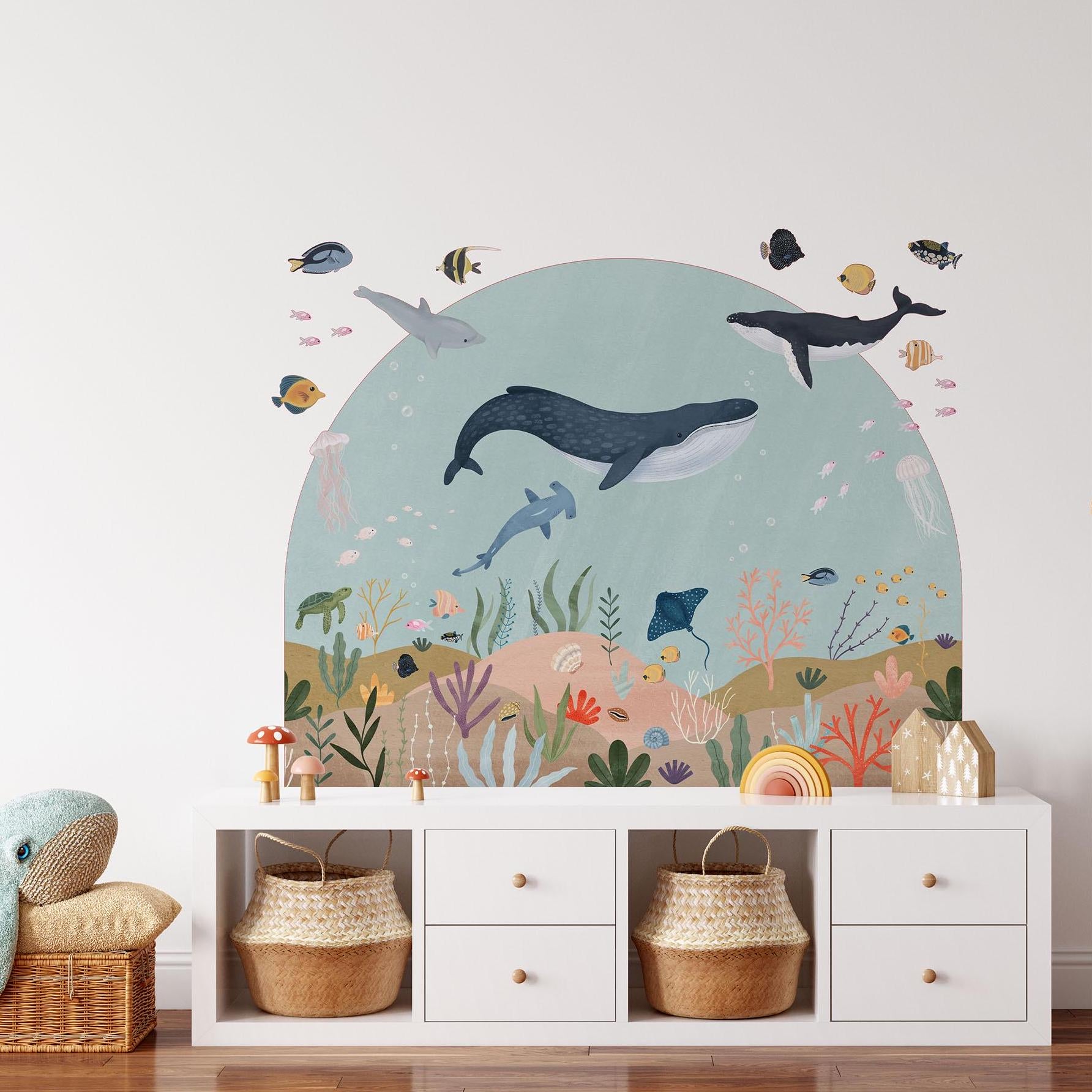The Sea Animals Arch Wall Sticker by My Store creates an enchanting underwater scene with whales, dolphins, and vibrant fish. Positioned above a white dresser decorated with baskets and a plush toy, it adds playful decor to the room.