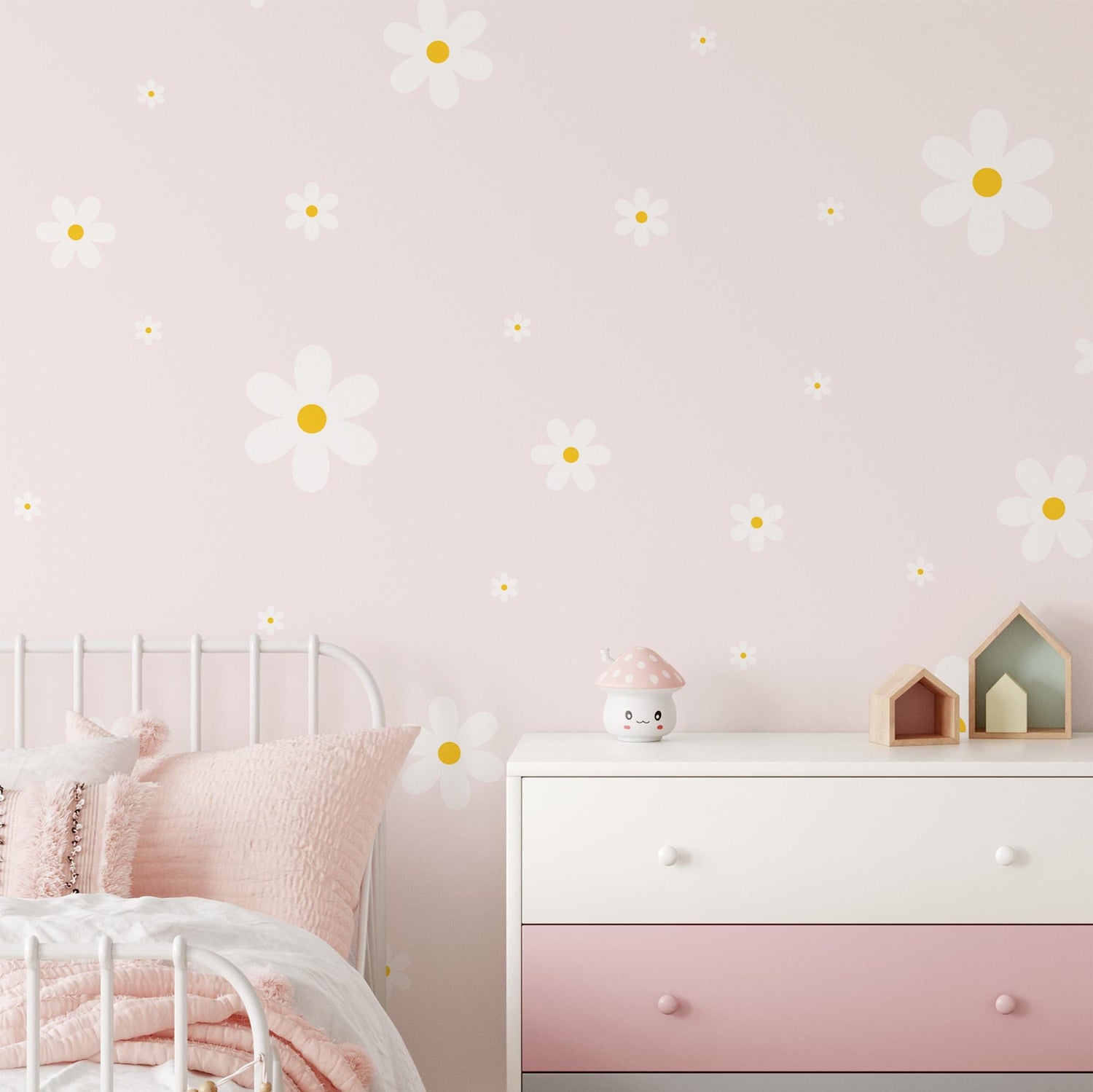 A pastel nursery features a white crib with a pink pillow. A pink and white dresser displays a mushroom-shaped nightlight and wooden house decor, while My Stores Daisy Wall Decals bring charm to the walls.