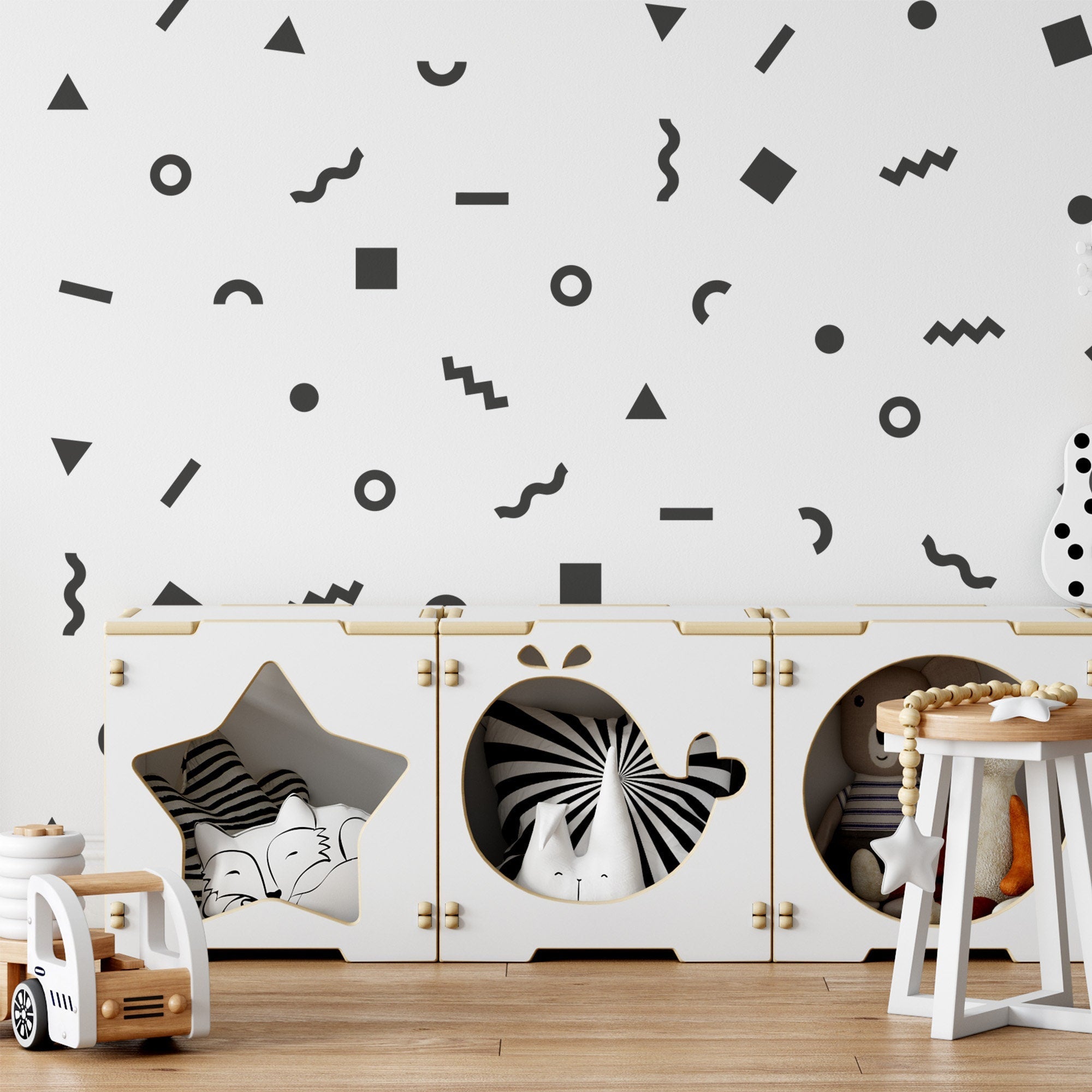 A childrens room with geometric walls includes white modular storage cubes filled with toys, a small wooden chair holding a stuffed animal, and a white toy vehicle. The modern monochrome decor is playful and enhanced by My Stores Retro Doodle Stickers, adding vintage charm.