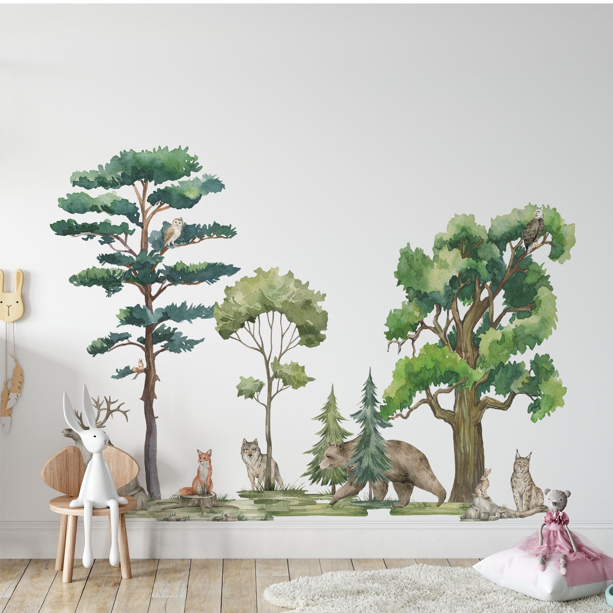 A childrens room boasts a wall mural of forest scenes featuring a fox, wolf, bear, and owl. My Stores Woodland Animals Nursery Stickers add charm with a plush deer on a wooden chair and a soft toy rabbit resting near a pillow on the floor.