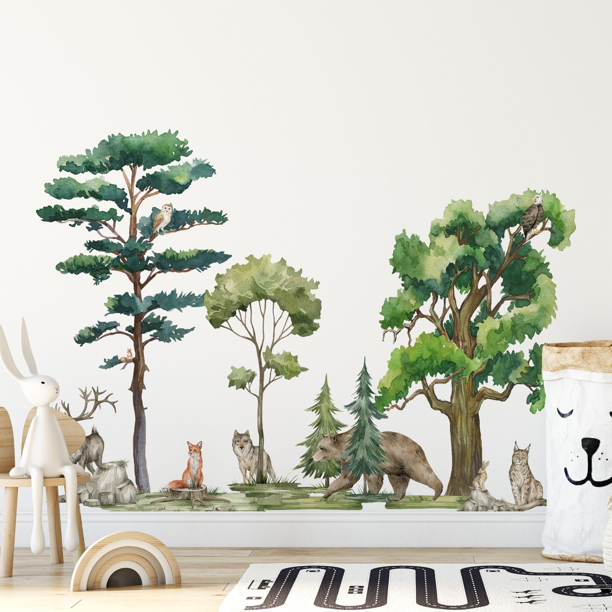 A nursery room features a whimsical forest-themed mural adorned with My Stores Woodland Animals Nursery Stickers, including a bear, fox, and owl among trees. A wooden bunny chair and a pillow with a sleeping face rest on the floor, beside a partially visible road-printed rug.