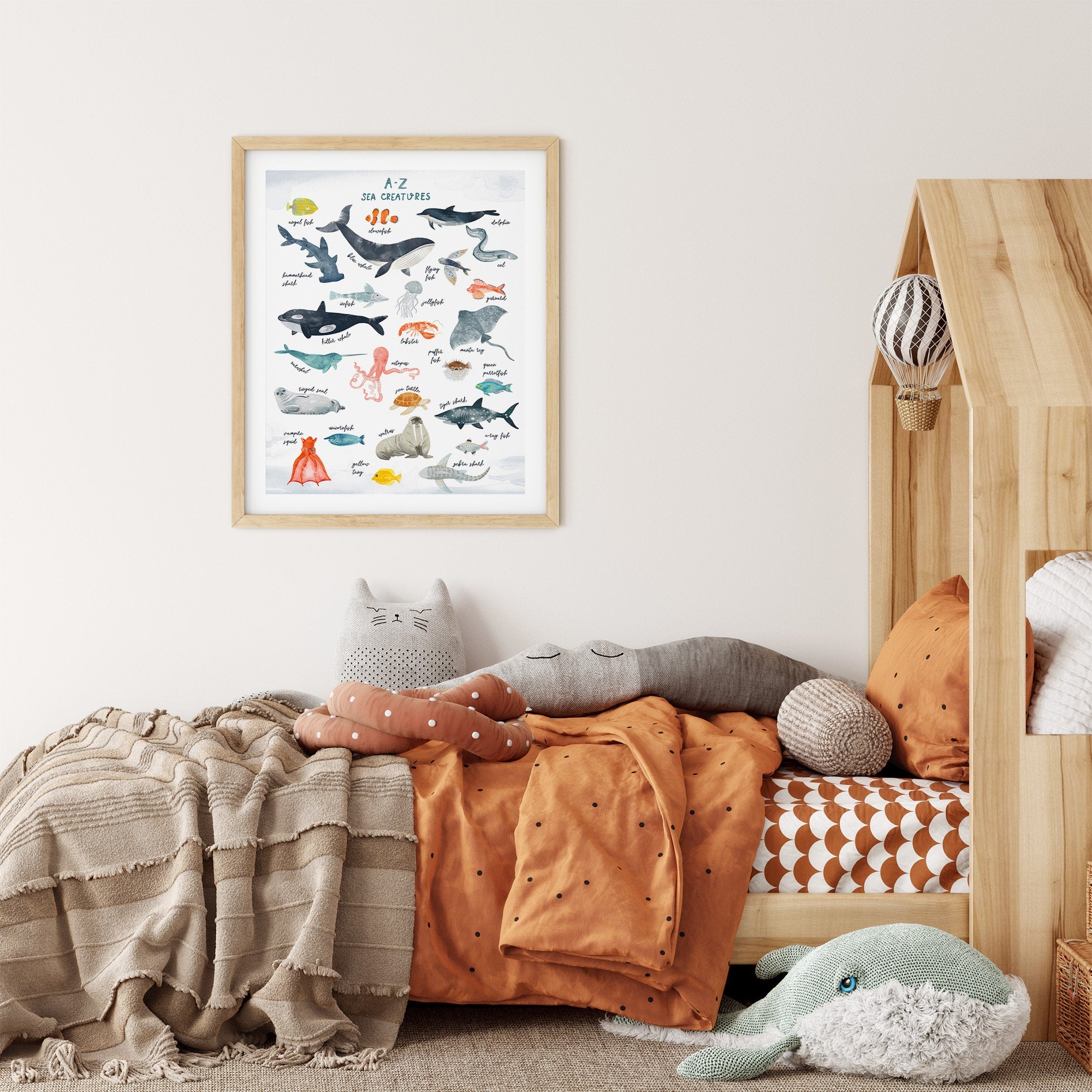 A cozy childrens bedroom features a house-shaped wooden bed with orange bedding, adorned with plush toys like a whale and cat. Above the bed hangs the A-Z Sea Creatures Nursery Print by My Store, showcasing various sea creatures.