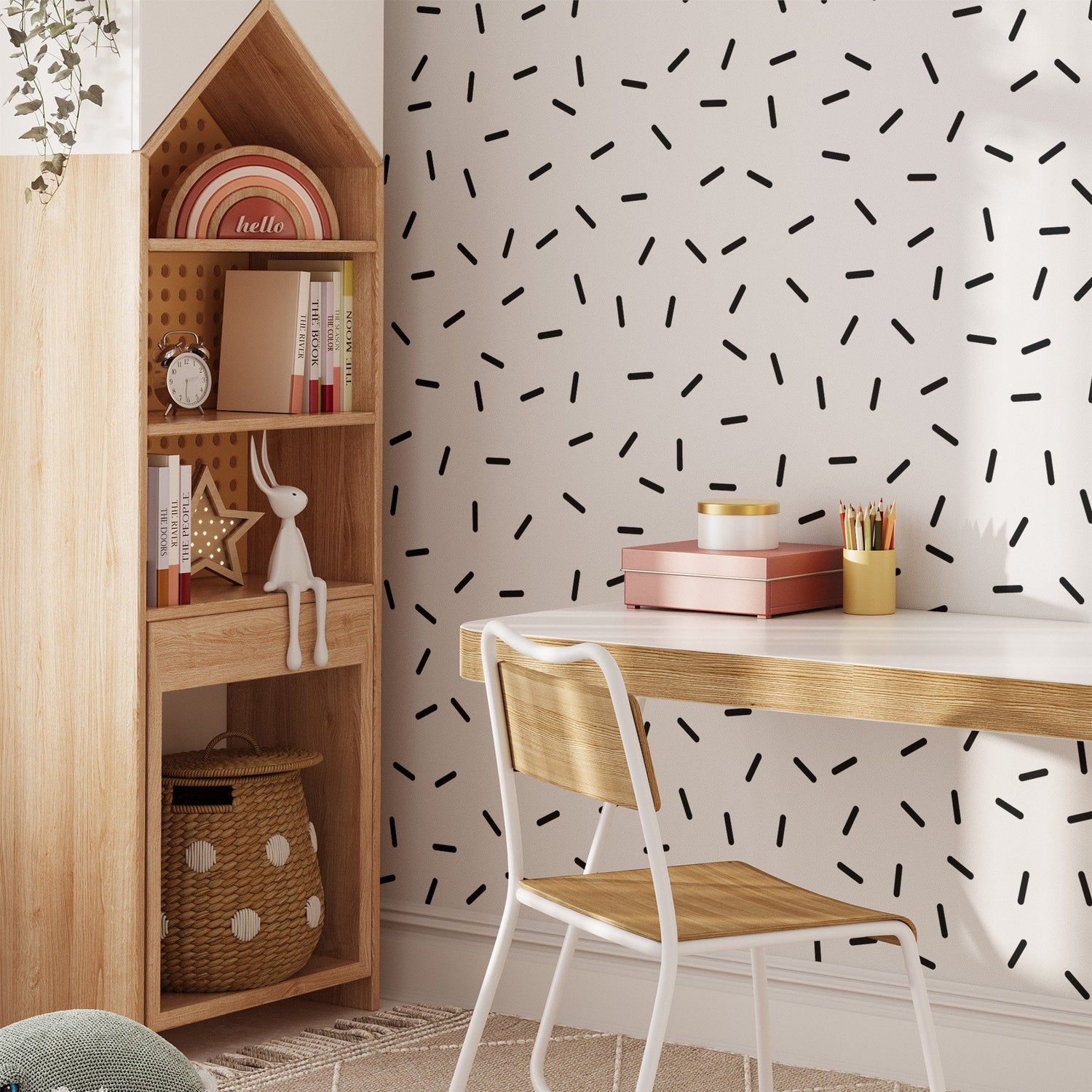 A minimalist workspace includes a wooden desk and chair, with a white wall decorated using My Stores Black Sprinkle Stickers. A wooden shelf beside the desk holds books, decorative items, and a toy rabbit, while a woven basket rests on the floor.