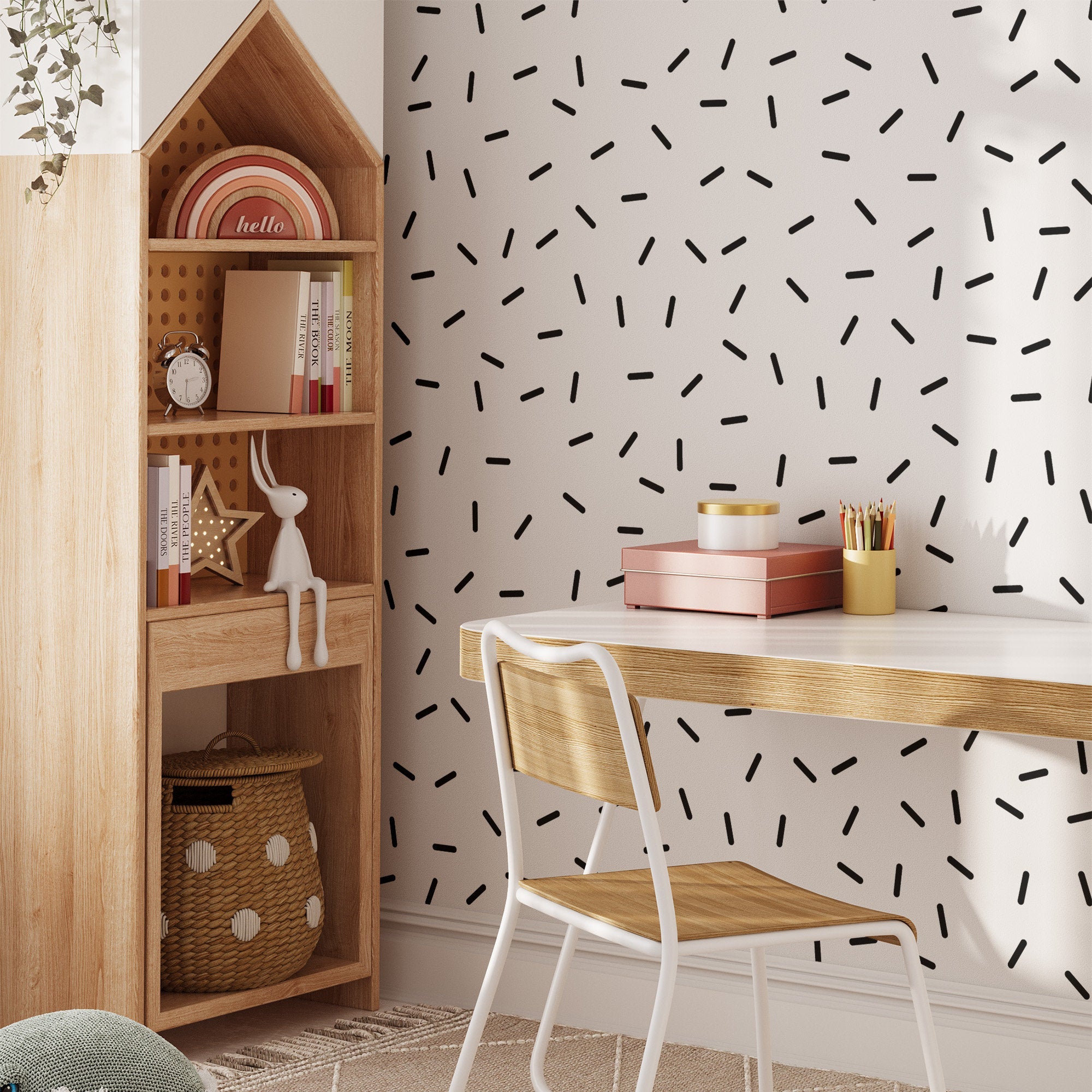 A minimalist workspace includes a wooden desk and chair, with a white wall decorated using My Stores Black Sprinkle Stickers. A wooden shelf beside the desk holds books, decorative items, and a toy rabbit, while a woven basket rests on the floor.