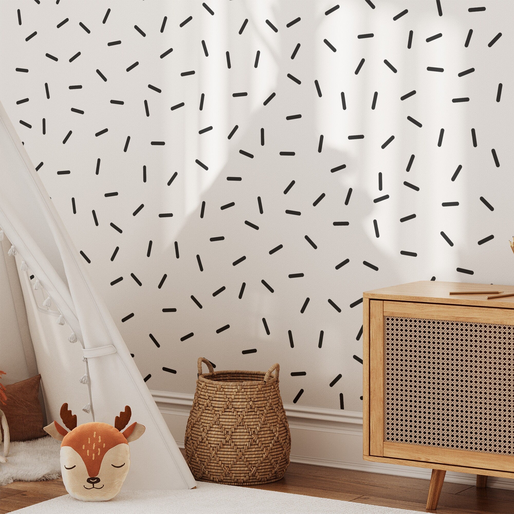 A cozy childrens room features playful elements: a triangular white tent beside a wooden cabinet with textured doors, a deer plush toy on the floor, and a woven basket nearby. The wall is adorned with scattered black lines using My Stores Black Sprinkle Stickers, which are removable nursery decor decals.