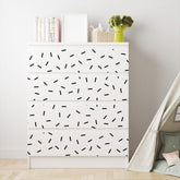 A charming nursery features a white dresser with black geometric designs next to a small teepee tent. The dresser displays books, frames, a woven basket, and a plush broccoli toy. Black Sprinkle Stickers by My Store add playfulness to the rooms airy and minimalist decor.