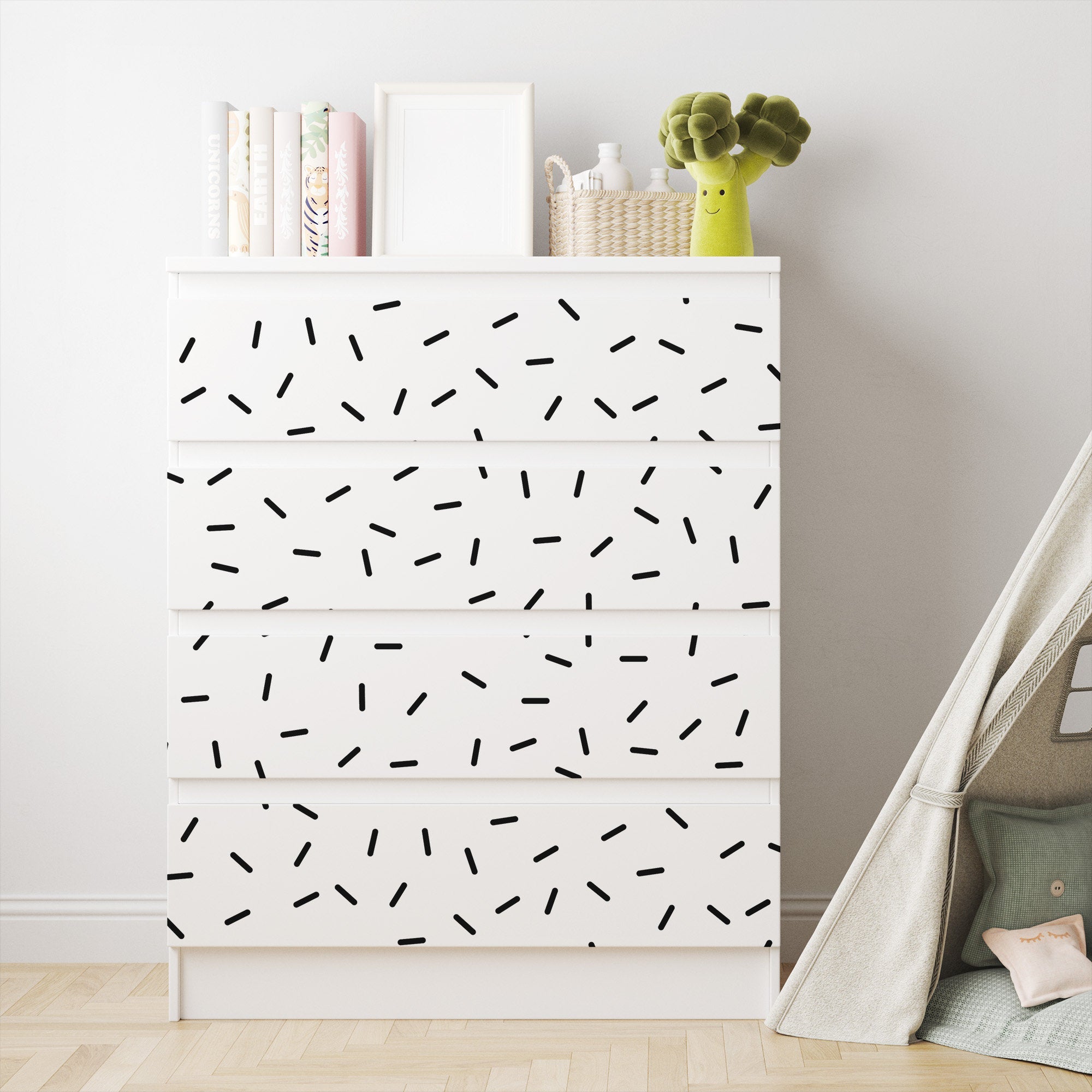 A charming nursery features a white dresser with black geometric designs next to a small teepee tent. The dresser displays books, frames, a woven basket, and a plush broccoli toy. Black Sprinkle Stickers by My Store add playfulness to the rooms airy and minimalist decor.