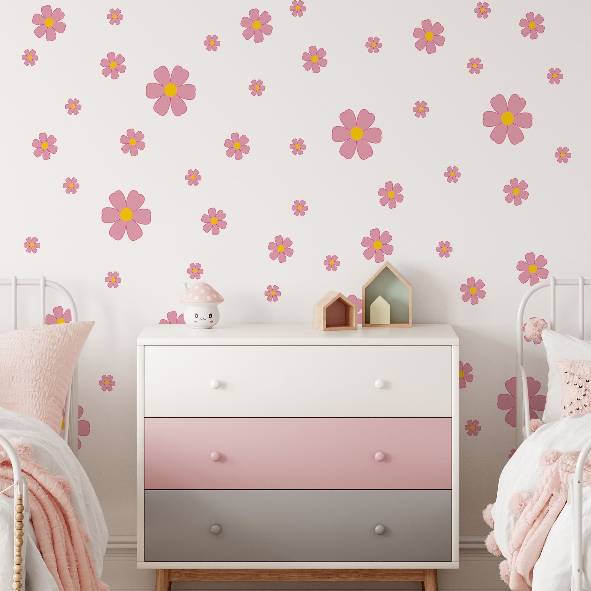 A childrens room with a white dresser featuring pink and gray drawers is decorated with My Stores Pink Daisy Stickers. The wall has removable decals alongside yellow floral designs, while pastel bedding and small decorative house models on the dresser add whimsy.