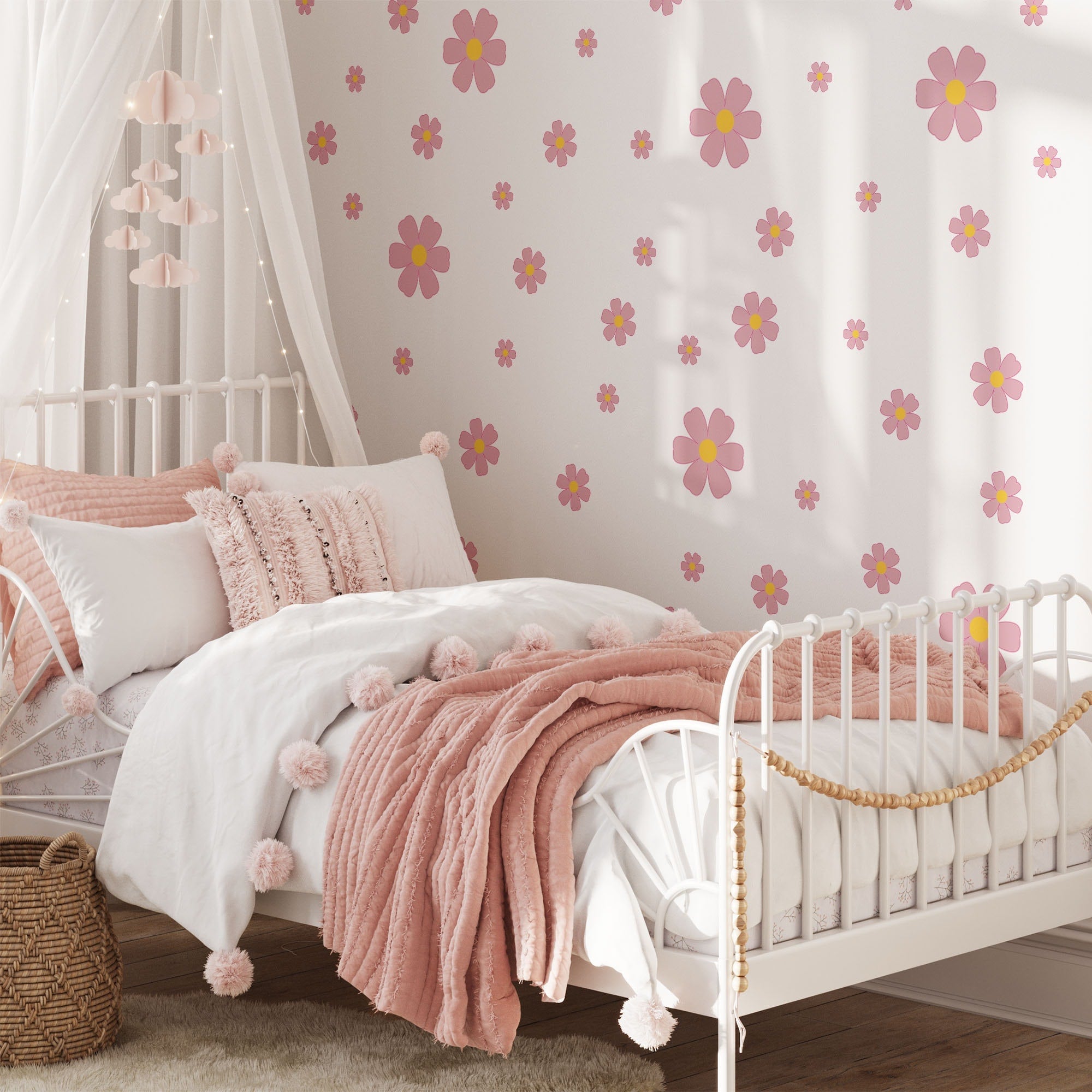 A cozy bedroom boasts a white metal bed frame with pink and white pillows, a fluffy blanket, and a canopy above. The wall is decorated with My Stores Pink Daisy Stickers. A woven basket sits by the bed, while soft light filters through the window.