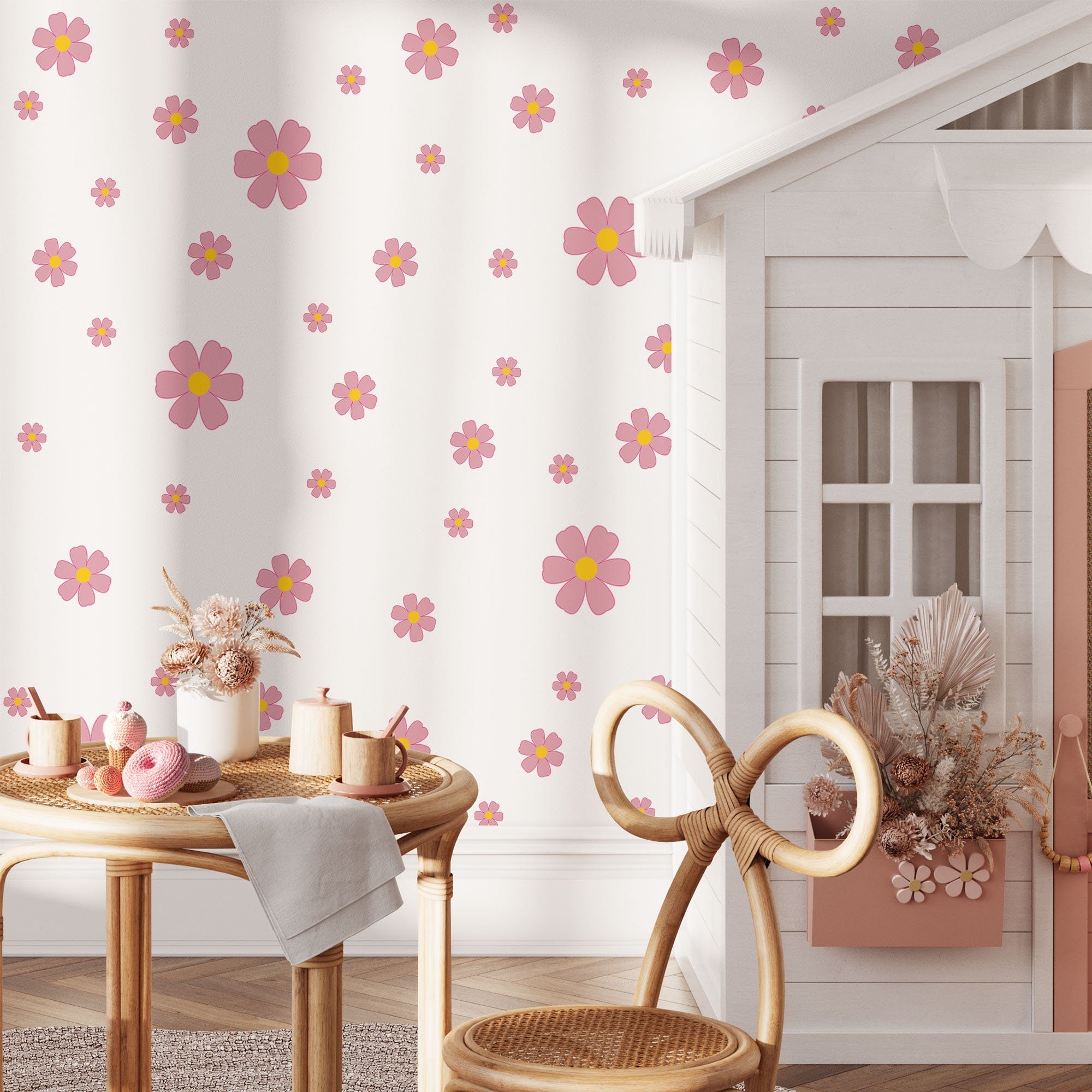 A cozy playroom features a small white playhouse and a table set with toys. My Stores Pink Daisy Stickers add charm to the wall. A wooden chair stands in the foreground, complemented by artfully arranged dried flowers on the table.