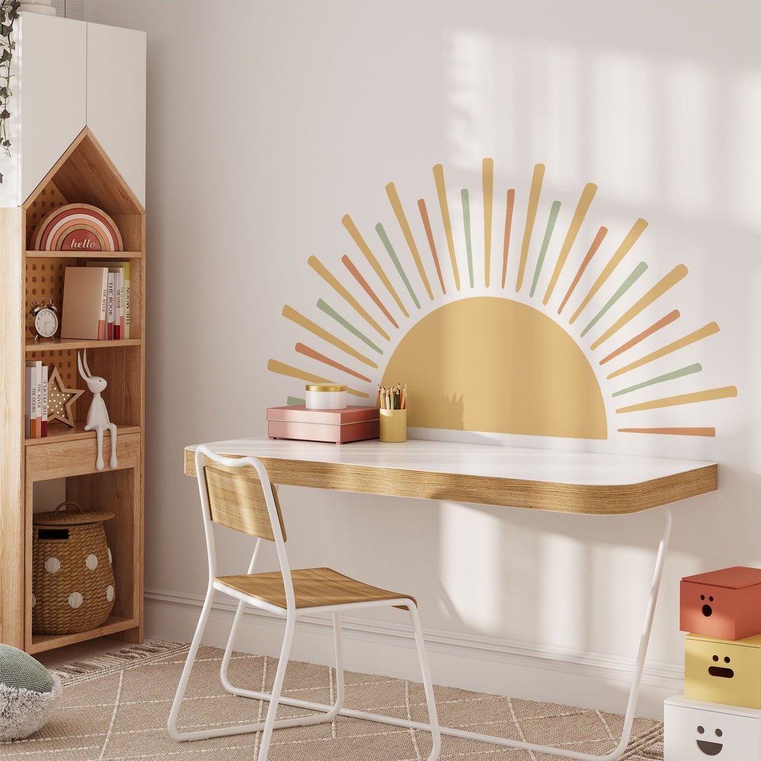 A modern study area with a minimalist white desk with wooden accents, chair, and colorful storage boxes. The Boho Sun Sticker from My Store adds warmth to the wall. A house-shaped bookshelf adds charm, holding books and a small plant.