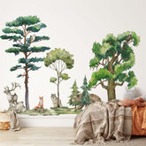 A cozy room showcases a nature-themed wall mural adorned with My Stores Woodland Animals Nursery Stickers, featuring trees and animals like deer, foxes, wolves, bears, and owls. A bed with pillows and blankets sits in the foreground, creating a warm, inviting atmosphere.