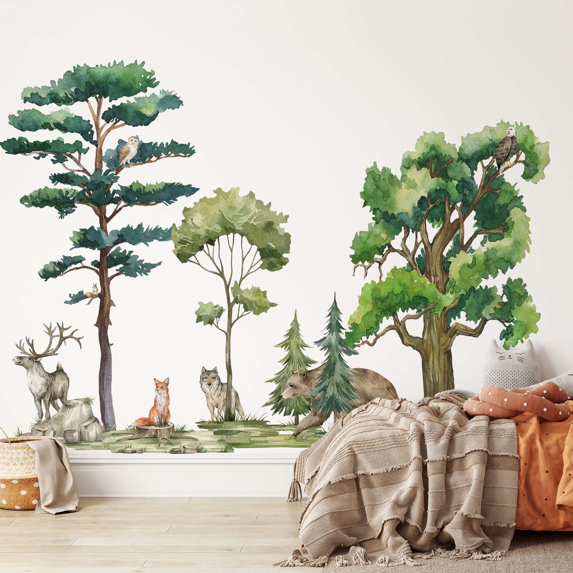A cozy room showcases a nature-themed wall mural adorned with My Stores Woodland Animals Nursery Stickers, featuring trees and animals like deer, foxes, wolves, bears, and owls. A bed with pillows and blankets sits in the foreground, creating a warm, inviting atmosphere.