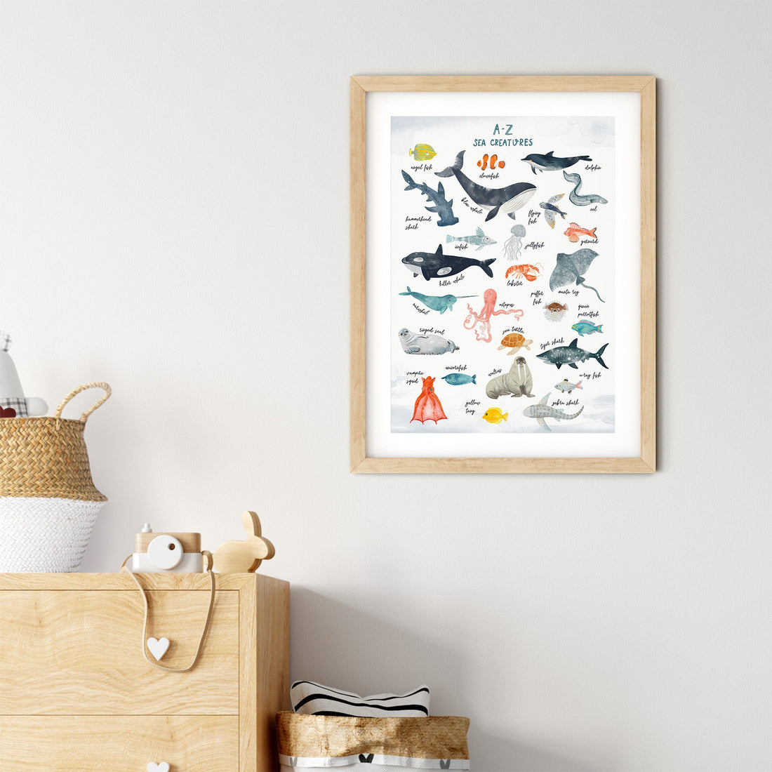 My Stores A-Z Sea Creatures Nursery Print, framed and displayed on the wall, features illustrations of sea creatures labeled alphabetically. Below, a wooden dresser holds decorative items like a woven basket and small white bottle, creating a charming nursery art atmosphere.