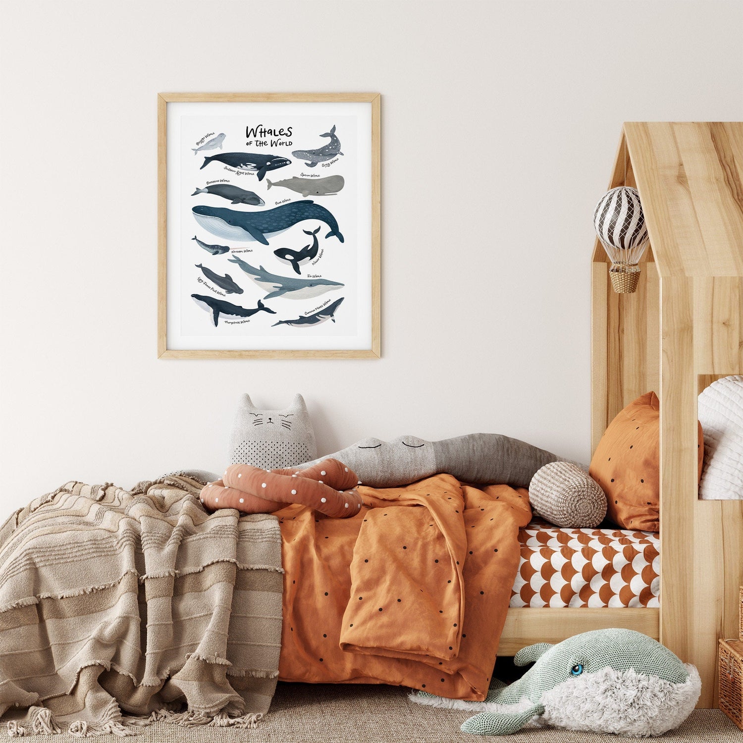 A cozy childrens room includes a wooden bed with animal-shaped cushions like a cat and whale. The orange and beige bedding complements My Stores Whales Of The World Nursery Print, featuring various whale illustrations as nursery art.